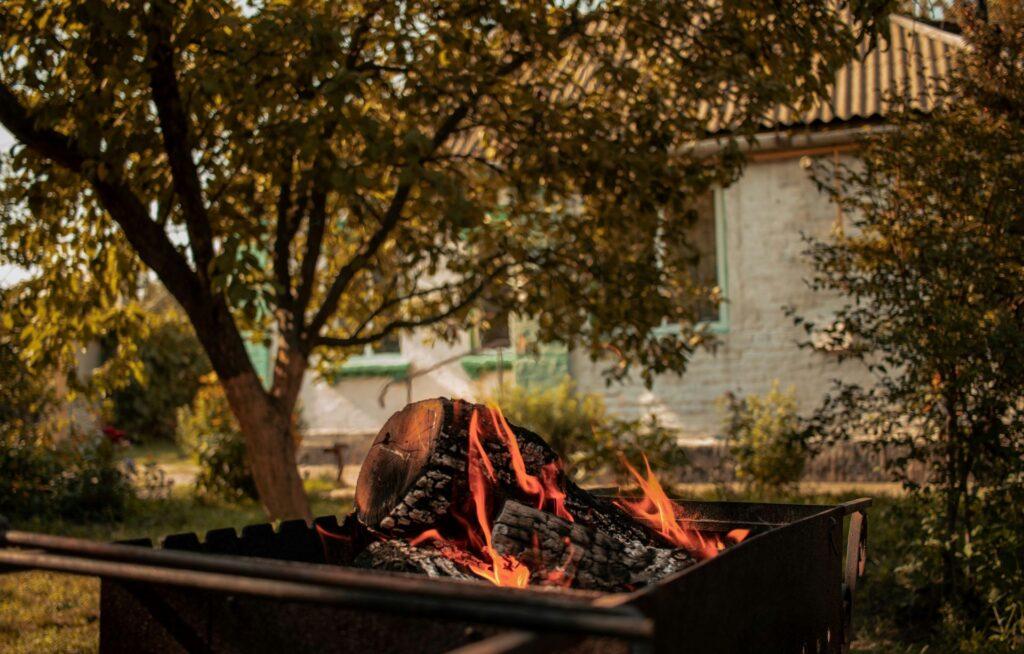 Feuerstelle im Garten mit glühendem Holz, im Hintergrund ein Baum und ein Haus in natürlicher Umgebung.