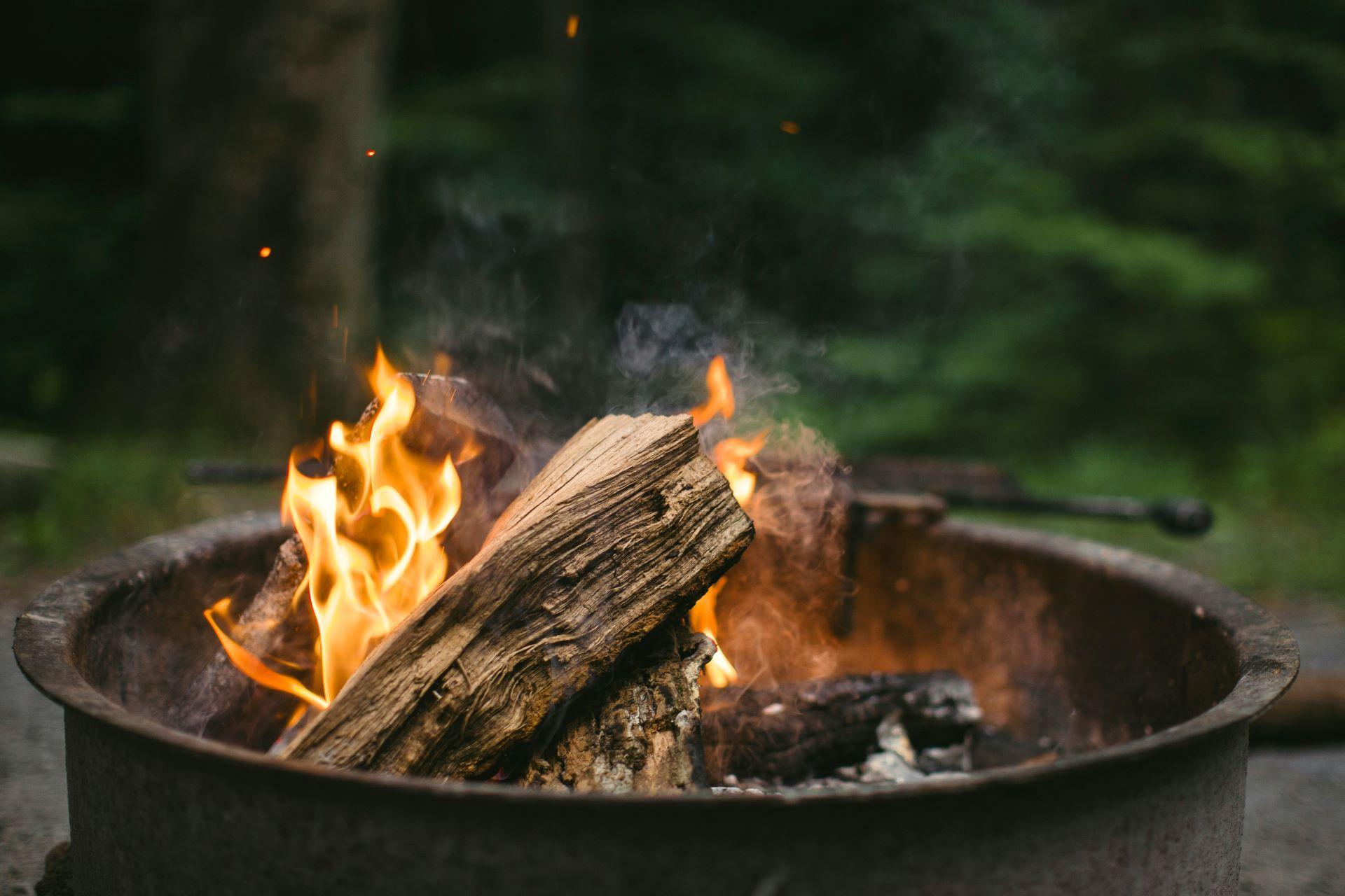 Eine Feuerplatte im Freien mit brennendem Holz, Rauch steigt auf, umgeben von einer grünen Waldkulisse.