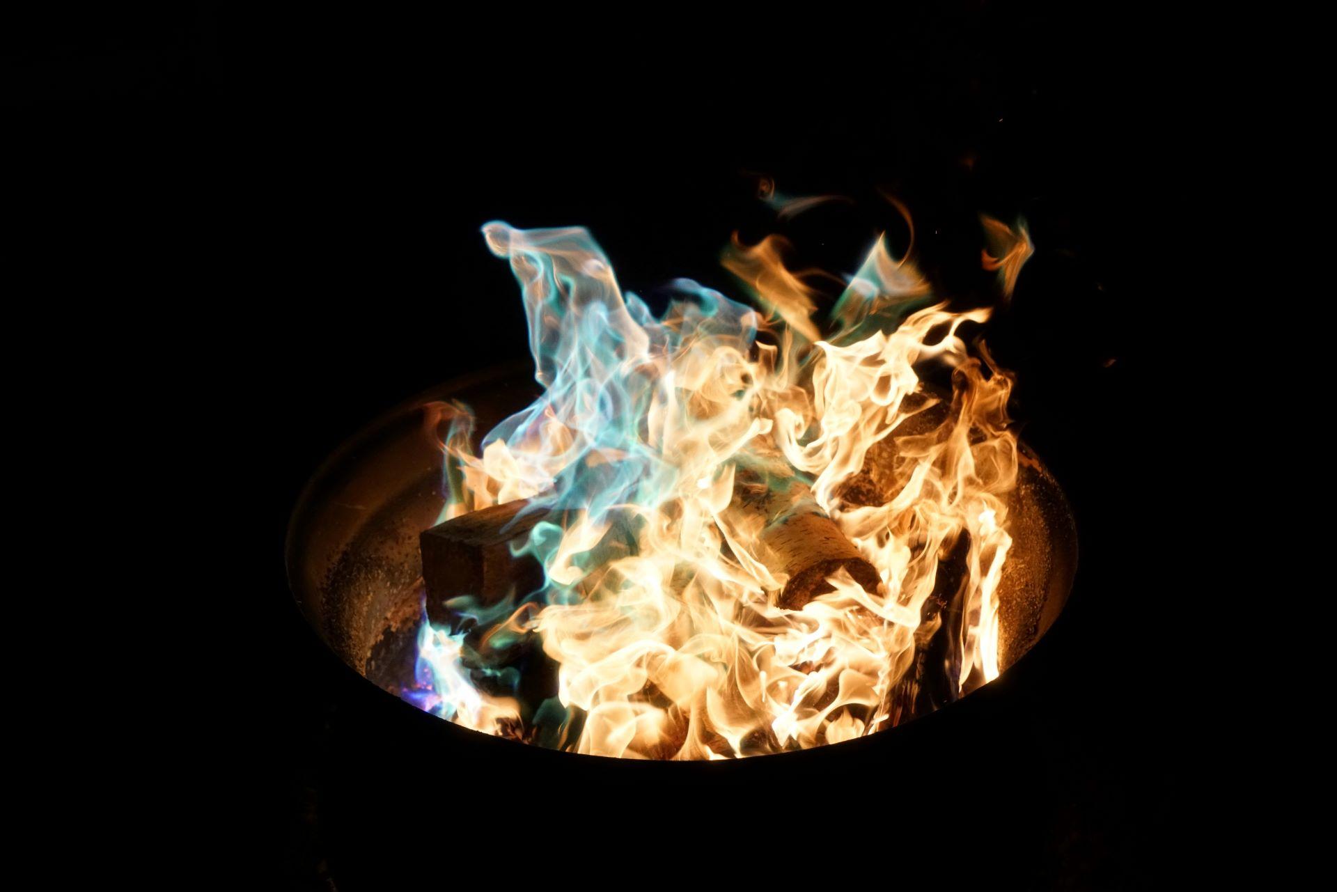 Nahaufnahme einer Feuerplatte mit bunten Flammen in der Dunkelheit, umgeben von Holzscheiten.