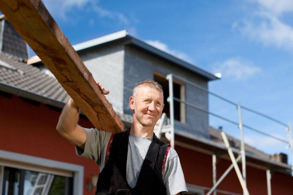 Dachsanierung durch erfahrenen Handwerker mit Holztragbalken vor Haus mit Geruest im Hintergrund